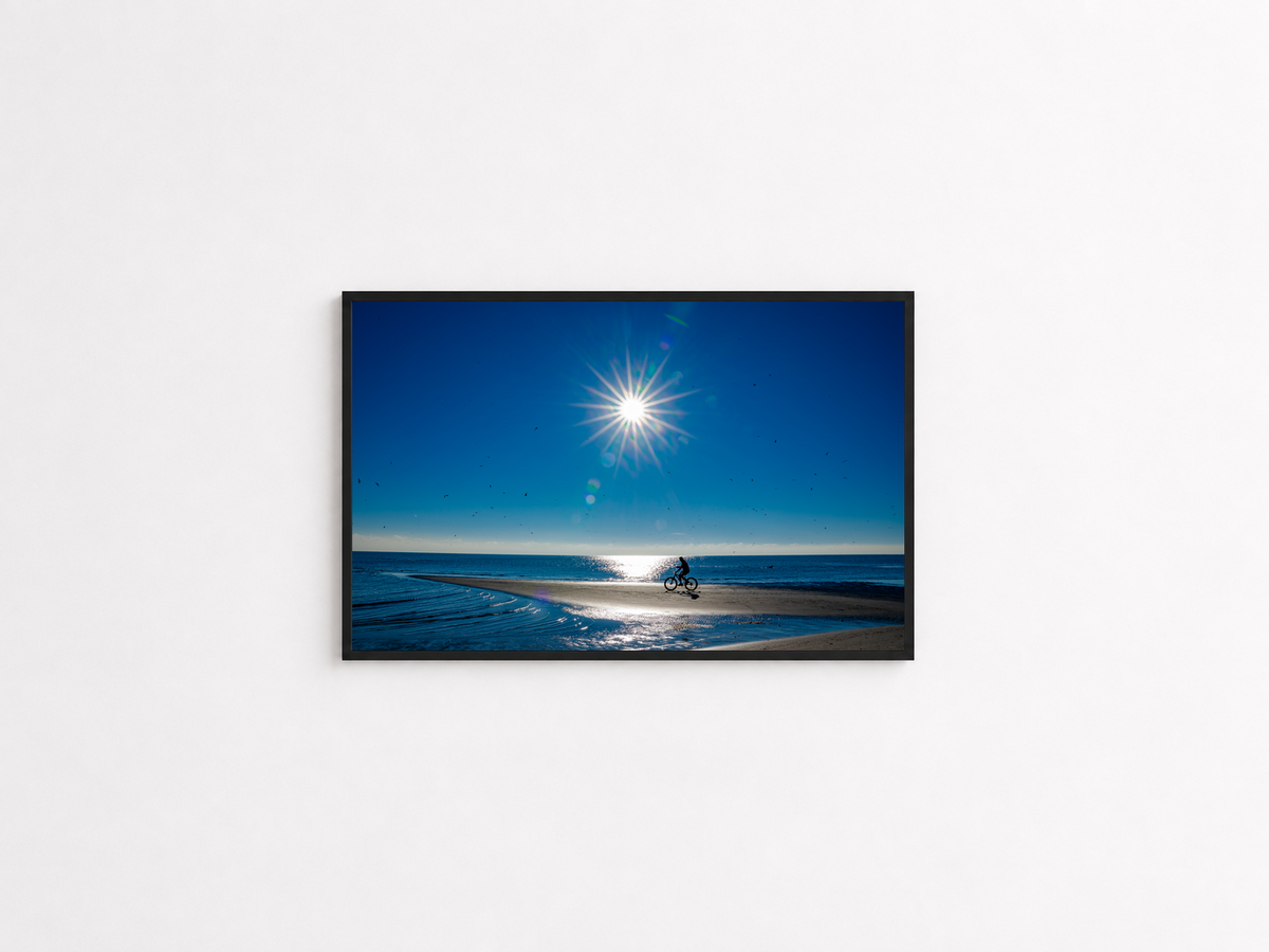 Cyclist crossing a sunlit coastal sandbar photography print in a black frame