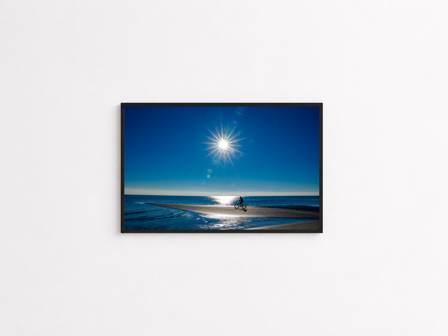 Cyclist crossing a sunlit coastal sandbar photography print in a black frame